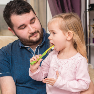 A father and daughter seated, a man and a young girl sitting, supervising teeth cleaning. Dad with toddler practicing good brushing.